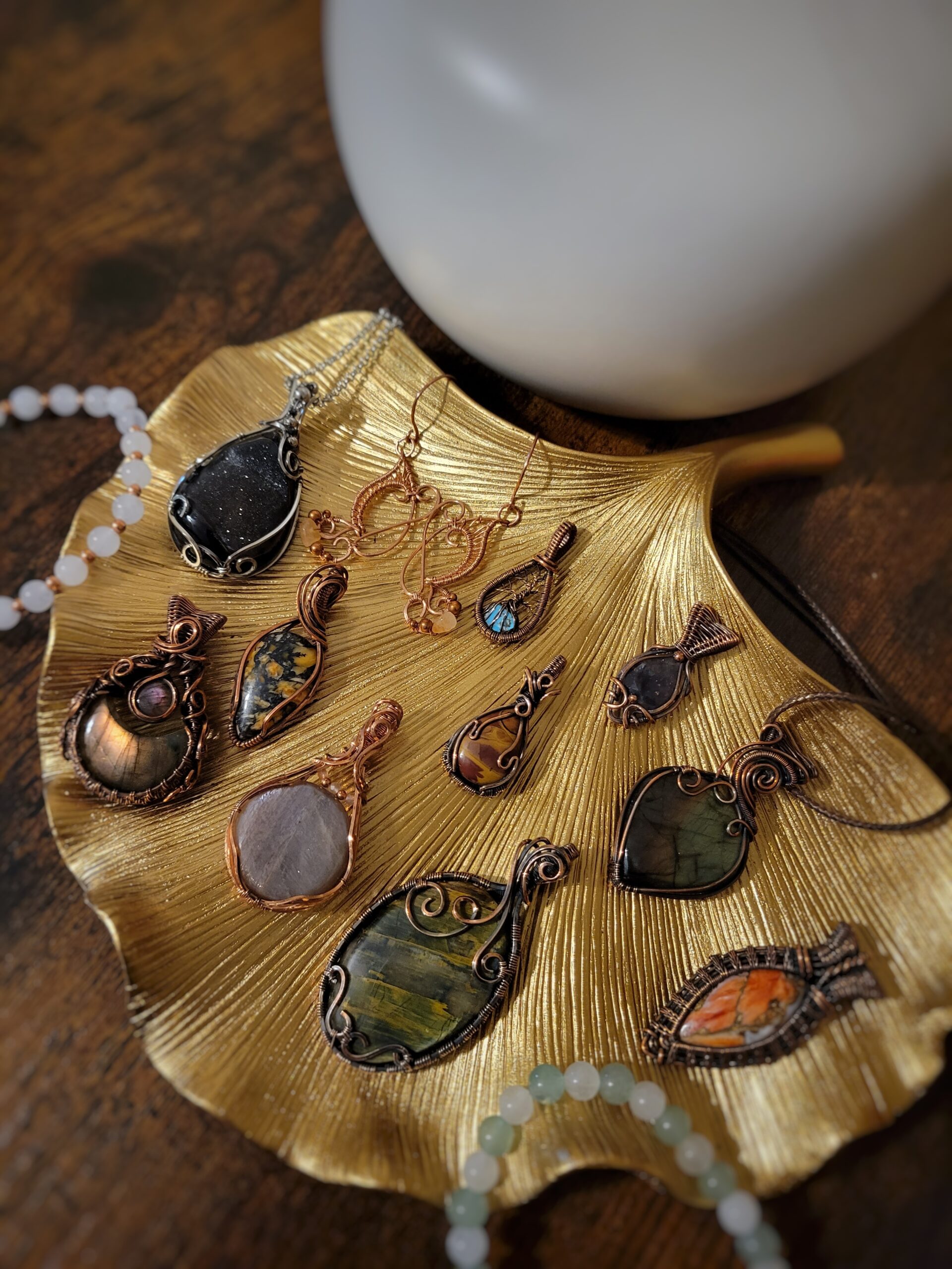An assortment of pendants and bracelets arranged on a gold, leaf-shaped tray. Pendants are wrapped in copper and sterling silver and the bracelets have stone and copper beads.