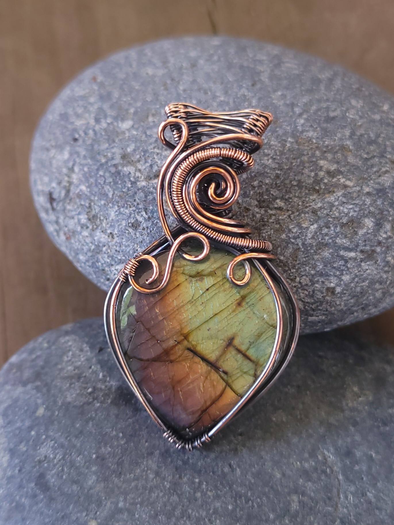 A finished rainbow labradorite pendant that was wrapped in copper wire and oxidized to look antique. It sits on two river stones with wooden boards in the background.